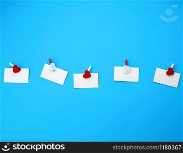 white empty paper rectangular business cards on decorative clothespins with a red heart lie on a blue background, place for an inscription
