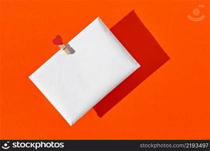 White envelope with a red heart on a clothespin, on an orange background. Love letter or Congratulations on Valentine?s Day, Mother?s Day, Women?s Day. View from above