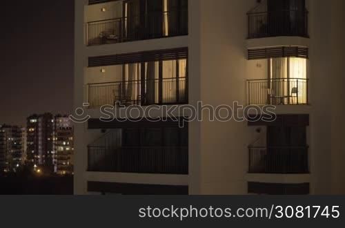 White facade of a multistory hotel looking building. It is night time and the sky is dark so the only visible light is pouring from the hotel windows. On the background there are some similar buildings