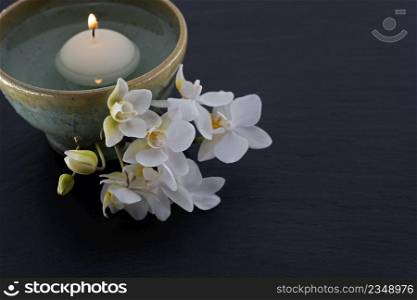 white floating candle in handmade ceramic bowl with a sprig of orchids on black slate board