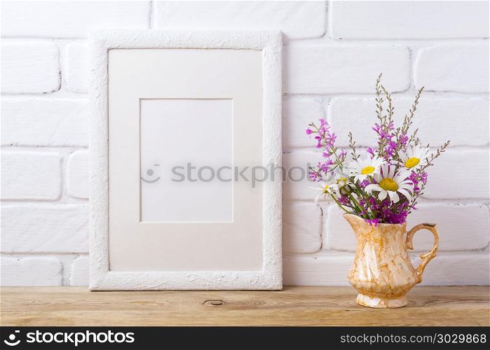 White frame mockup with chamomile and purple flowers in golden p. White picture frame with mat mockup with white chamomile and purple field flowers in golden pitcher vase. Empty frame mock up for presentation artwork. Template framing for modern art.