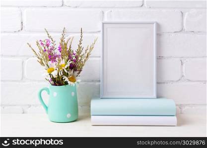White frame mockup with chamomile and purple flowers in mint gre. White frame mockup with white chamomile and purple field flowers in mint green pitcher vase and books. Empty frame mock up for presentation artwork. Template framing for modern art.