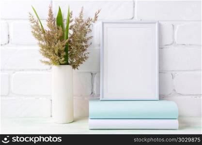 White frame mockup with grass and green leaves in cylinder vase. White frame mockup with meadow grass and green leaves in cylinder vase and books near painted brick wall. Empty frame mock up for presentation artwork. Template framing for modern art.