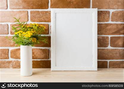 White frame mockup with wild rich golden yellow flowers in vase near exposed brick walls. Empty frame mock up for presentation design. Template framing for modern art.. White frame mockup with yellow flowers near exposed brick walls