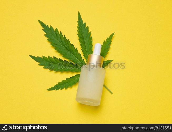 White glass bottle with pipette and green cannabis leaf on yellow background, top view