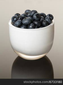 White glass cup full of blue blueberries reflecting on shiny gradient surface