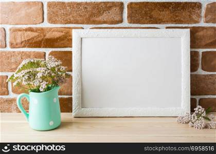 White landscape frame mockup with wild creamy pink flowers in mint pitcher near exposed brick wall. Empty frame mock up for presentation design. Template framing for modern art.. White landscape frame mockup creamy pink flowers in mint pitcher