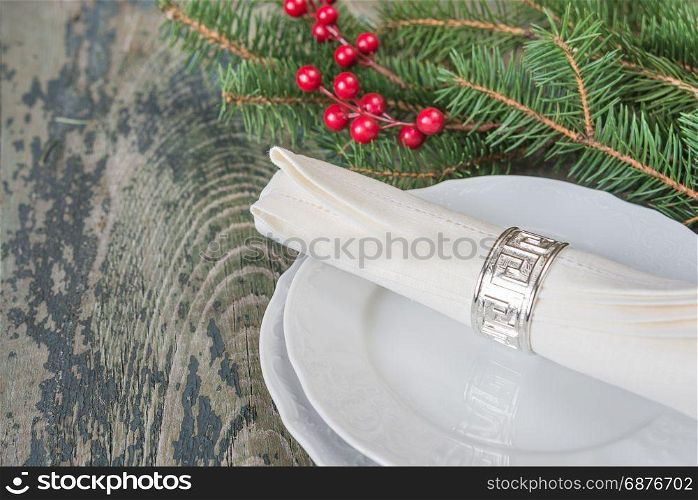 White linen napkin, silver napkin ring are on the white dinner plate, as well as red holly berries and green spruce branches which is located on a old wooden table, with space for text