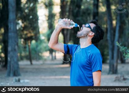 white man drinking water and listening to music in a park while exercising.