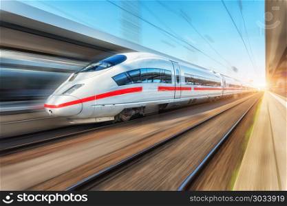 White modern high speed train in motion on railway station at sunset. Passenger train on railroad track with motion blur effect in Europe. Railway platform. Industrial landscape. Railway tourism. White modern high speed train in motion on railway station