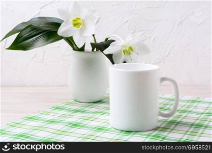 White mug mockup with lily in vase on green checkered napkin. Empty mug mock up for design promotion. . White mug mockup with lily in vase on green checkered napkin