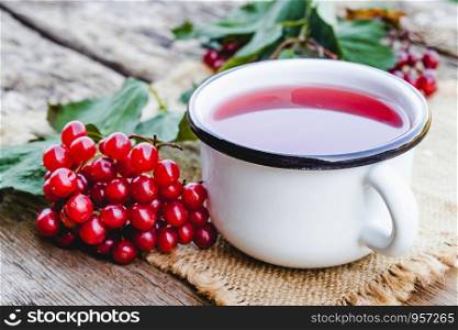 White mug or cup of hot viburnum tea on a wooden table next to red viburnum berries. Source of natural vitamins. Used in folk medicine. Close-up.. White mug or cup of hot viburnum tea on a wooden table next to red viburnum berries. Source of natural vitamins. Used in folk medicine.