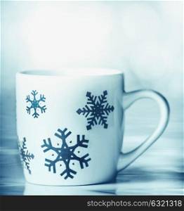White mug with blue snowflakes on blue table and bokeh background, front view. Happy winter holiday card layout