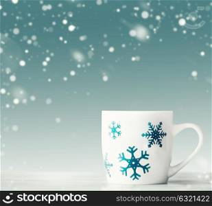 White mug with blue snowflakes on white table at blue background with snowfall, front view. Happy winter holiday card layout