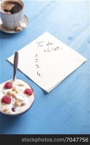 White napkin with a draft of a to do list, surrounded by a bowl of yogurt with cereal and raspberry and a mug of aromatic coffee, on a blue background.