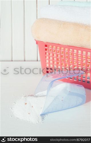 White, orange and blue fluffy bath towels in the red laundry basket and spilled washing powder in measuring cup on the background of white boards