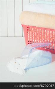 White, orange and blue fluffy bath towels in the red laundry basket and spilled washing powder in measuring cup on the background of white boards