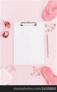 white paper clipboard surrounded with miniature luggage bag sunglasses paperclips pencil flip flops pink background