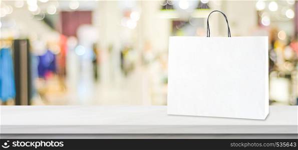 White paper shopping bag standing on white marble table over blurred store background with copy space, business marketing background, banner, mock up, template for retail, sale product