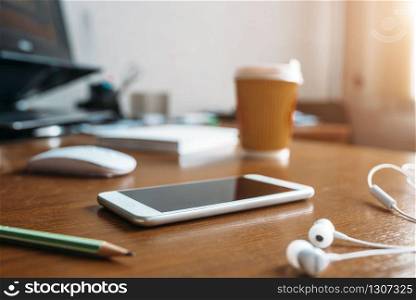 White phone, headphones, pc mouse and cup of coffee on wooden table in office closeup, nobody. Modern business workplace. White phone, headphones, mouse and cup of coffee