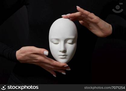 White plaster mask face is holding woman&rsquo;s fingers on a black background, copy space. Concept social psychological masks. Two woman&rsquo;s hands hold white gypsum mask face on a black background. Concept social psychological masks