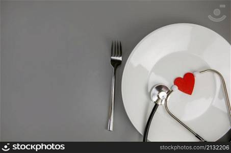 White plate with red heart and stethoscope and knife and fork on grey background with copy space,health care and diet concept space for text. White plate with red heart and stethoscope and knife and fork on grey background with copy space,health care and diet concept