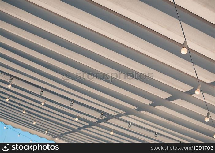 White roofing of a terrace in a street cafe or restaurant, with bulbs hanging under the awning, in the midday sun against blue sky