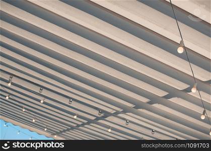 White roofing of a terrace in a street cafe or restaurant, with bulbs hanging under the awning, in the midday sun against blue sky
