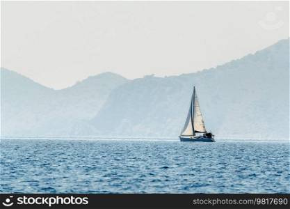 White sailing yacht surrounded by magnificent nature. High mountains and hills, blue sea surface, and a cloudless sky. A dreamy idyllic landscape.. Beautiful seascape with white sailboat