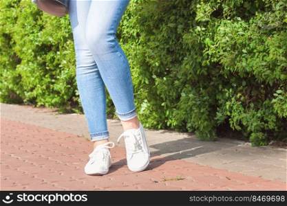 White sneakers on female legs in blue jeans on the asphalt and green plant background.. White sneakers on girl legs in blue jeans
