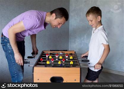 white t-shirt, foosball, dad and son, little boy, play football, twist levers, home games, white hair, smile. Cute boy playing table football with his emotional dad in the room