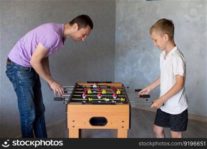 white t-shirt, foosball, dad and son, little boy, play football, twist levers, home games, white hair, smile. Cute boy playing table football with his emotional dad in the room
