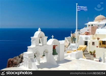 White tower with bells and the Greek flag on a background of blue sea and sky in the village of Oia. Santorini, Greece.