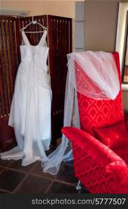 White wedding dress hanging on hanger behind a red chair on which is the vail.