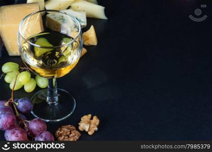 White wine, various types of cheese, walnuts and grapes on a black background of slate stone