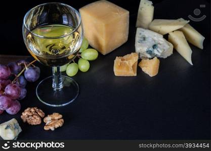 White wine, various types of cheese, walnuts and grapes on a black background