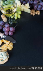 White wine, various types of cheese, walnuts and grapes on a black slate stone background