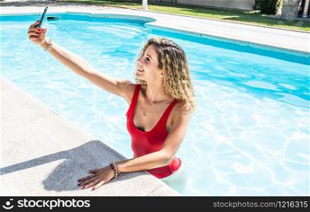 white woman in a red swimsuit taking a picture with her cell phone in the pool