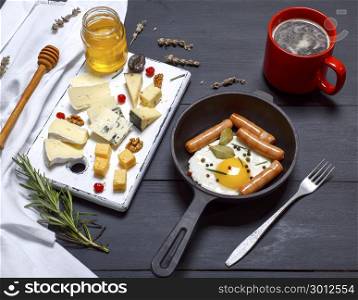 white wooden board with pieces of different kind of cheese and a round cast-iron frying pan with fried egg and sausages, top view