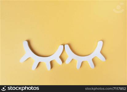 White wooden closed eyes with eyelashes on pastel yellow background. Stylish wall decor. Top view, flat lay. Concept of vivid sweet dreams. Accessories for kids room. Minimal style.