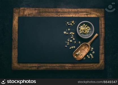 Whole dried chamomile flowers and wooden shovel spoon on dark chalkboard background, top view with copy space for your design. Healing herbs and herbal medicine concept. Horizontal