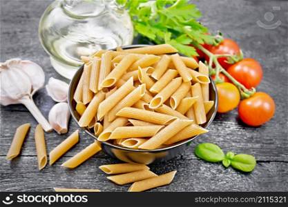Whole grain flour penne pasta in a bowl, tomatoes, garlic, vegetable oil in a decanter and parsley on black wooden board background