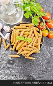Whole grain flour penne pasta in a bowl, tomatoes, garlic, vegetable oil in a decanter and parsley on dark wooden board background from above