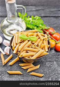Whole grain flour penne pasta in a bowl, tomatoes, garlic, vegetable oil in a glass decanter and parsley on dark wooden board background