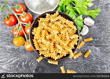 Whole grain wheat flour fusilli pasta in a bowl, tomatoes, garlic, vegetable oil in a decanter and parsley on dark wooden board background from above