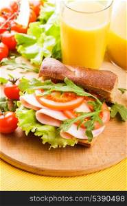 Wholegrain sandwich with ham, tomato, lattuce and arugula with glass of orange juice on the board
