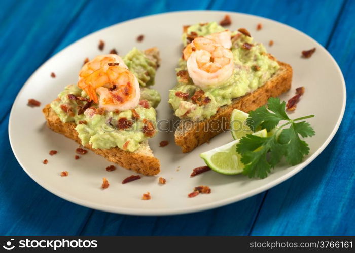Wholegrain toast bread slices with guacamole, fried shrimp and fried bacon pieces served on plate on blue wood (Selective Focus, Focus on the front of the shrimp on the left bread and the front of the right bread)
