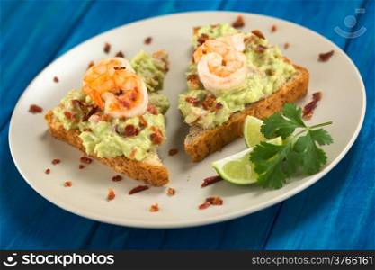 Wholegrain toast bread slices with guacamole, fried shrimp and fried bacon pieces served on plate on blue wood (Selective Focus, Focus on the front of the shrimp on the left bread and the front of the right bread)