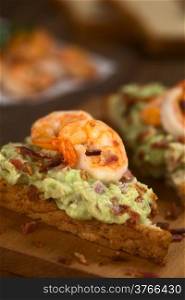 Wholegrain toast bread slices with guacamole, fried shrimp and fried bacon pieces on wooden board (Selective Focus, Focus on the front of the shrimp on the bread)