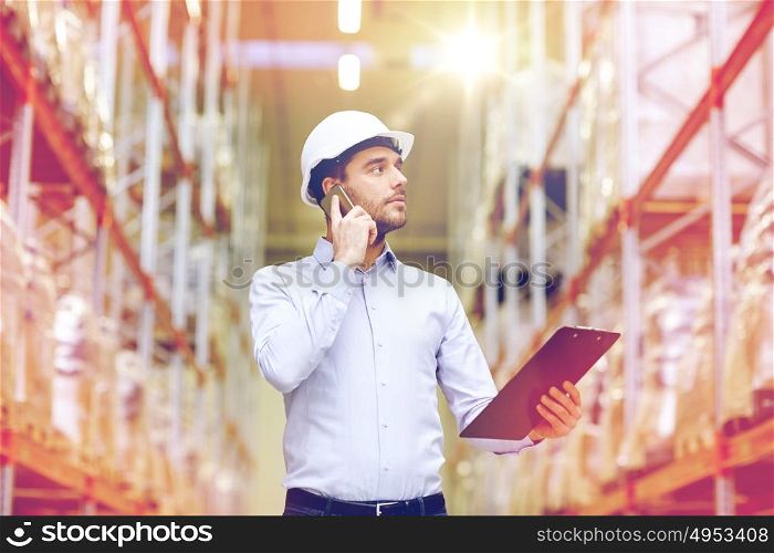 wholesale, logistic, business, export and people concept - businessman with clipboard calling on smartphone at warehouse. man with clipboard and smartphone at warehouse